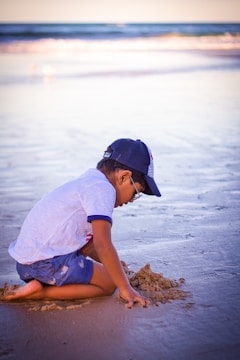 Sun-hatted toddler building a sandcastle on a sunny beach with clear blue waves in the background.