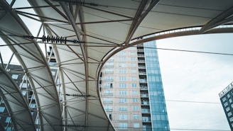 An upward view of a structure with geometric patterns created by metal beams and a fabric canopy. In the background, a modern high-rise building with glass windows is visible, creating a contrast between the architectural elements and the sky.