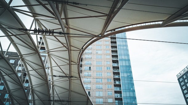 An upward view of a structure with geometric patterns created by metal beams and a fabric canopy. In the background, a modern high-rise building with glass windows is visible, creating a contrast between the architectural elements and the sky.