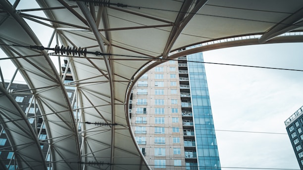 An upward view of a structure with geometric patterns created by metal beams and a fabric canopy. In the background, a modern high-rise building with glass windows is visible, creating a contrast between the architectural elements and the sky.