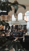 A spacious indoor bar area with high ceilings and arched windows, filled with people sitting and conversing at tables. The space is elegantly decorated with large chandeliers and vintage-style lighting. Lush green plants are partially visible in the foreground, adding a touch of nature to the sophisticated ambiance.