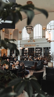 A spacious indoor bar area with high ceilings and arched windows, filled with people sitting and conversing at tables. The space is elegantly decorated with large chandeliers and vintage-style lighting. Lush green plants are partially visible in the foreground, adding a touch of nature to the sophisticated ambiance.