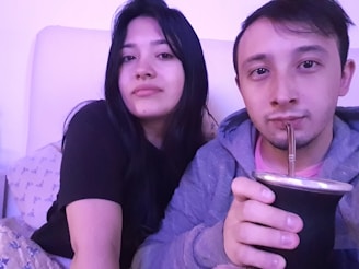 A man and a woman sit closely together, with the man holding a traditional mate cup with a metal straw. They are both looking at the camera, and the lighting is soft with a hint of a purple hue. They appear relaxed and comfortable, sitting on a couch with pillows.