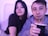 A man and a woman sit closely together, with the man holding a traditional mate cup with a metal straw. They are both looking at the camera, and the lighting is soft with a hint of a purple hue. They appear relaxed and comfortable, sitting on a couch with pillows.