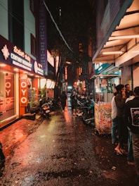 A gritty urban alleyway at dusk with a small group sharing a meal and conversation around a fire barrel.