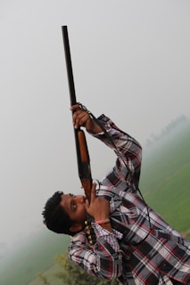 A person in a plaid shirt holding a shotgun pointed upwards, standing in an outdoor setting with a misty landscape and green fields in the background.