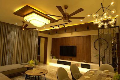 A modern living room with warm lighting features a ceiling fan, chandelier, and a rectangular ceiling light. The room includes a large curtain, a flat-screen TV mounted on a wooden panel, and several plants. The furniture consists of a sectional sofa, round coffee tables, and a dining table set for four.