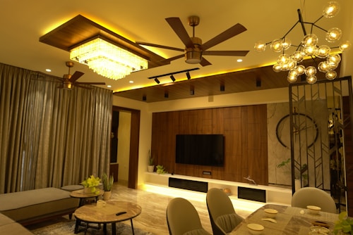 A modern living room with warm lighting features a ceiling fan, chandelier, and a rectangular ceiling light. The room includes a large curtain, a flat-screen TV mounted on a wooden panel, and several plants. The furniture consists of a sectional sofa, round coffee tables, and a dining table set for four.