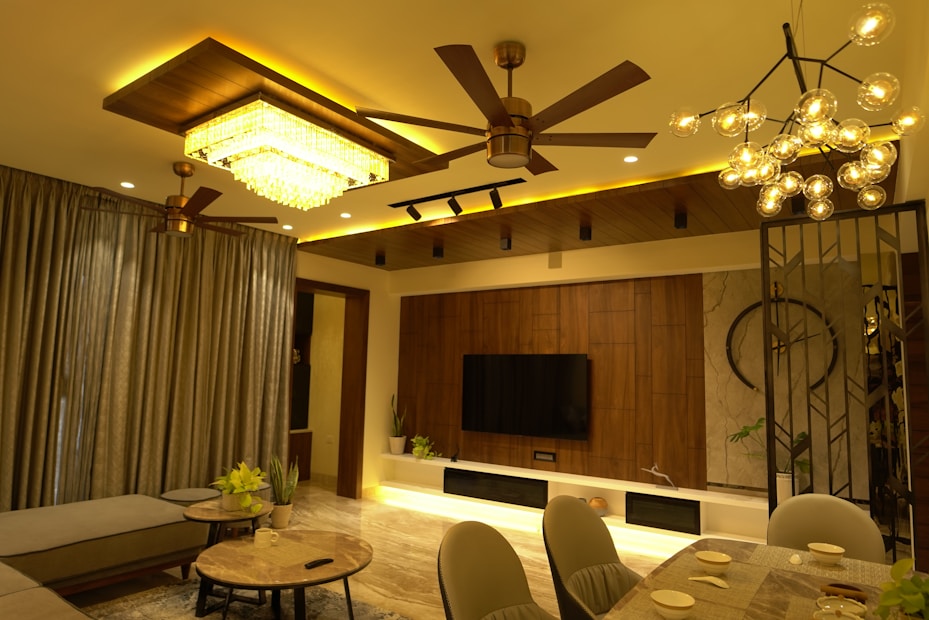 A modern living room with warm lighting features a ceiling fan, chandelier, and a rectangular ceiling light. The room includes a large curtain, a flat-screen TV mounted on a wooden panel, and several plants. The furniture consists of a sectional sofa, round coffee tables, and a dining table set for four.