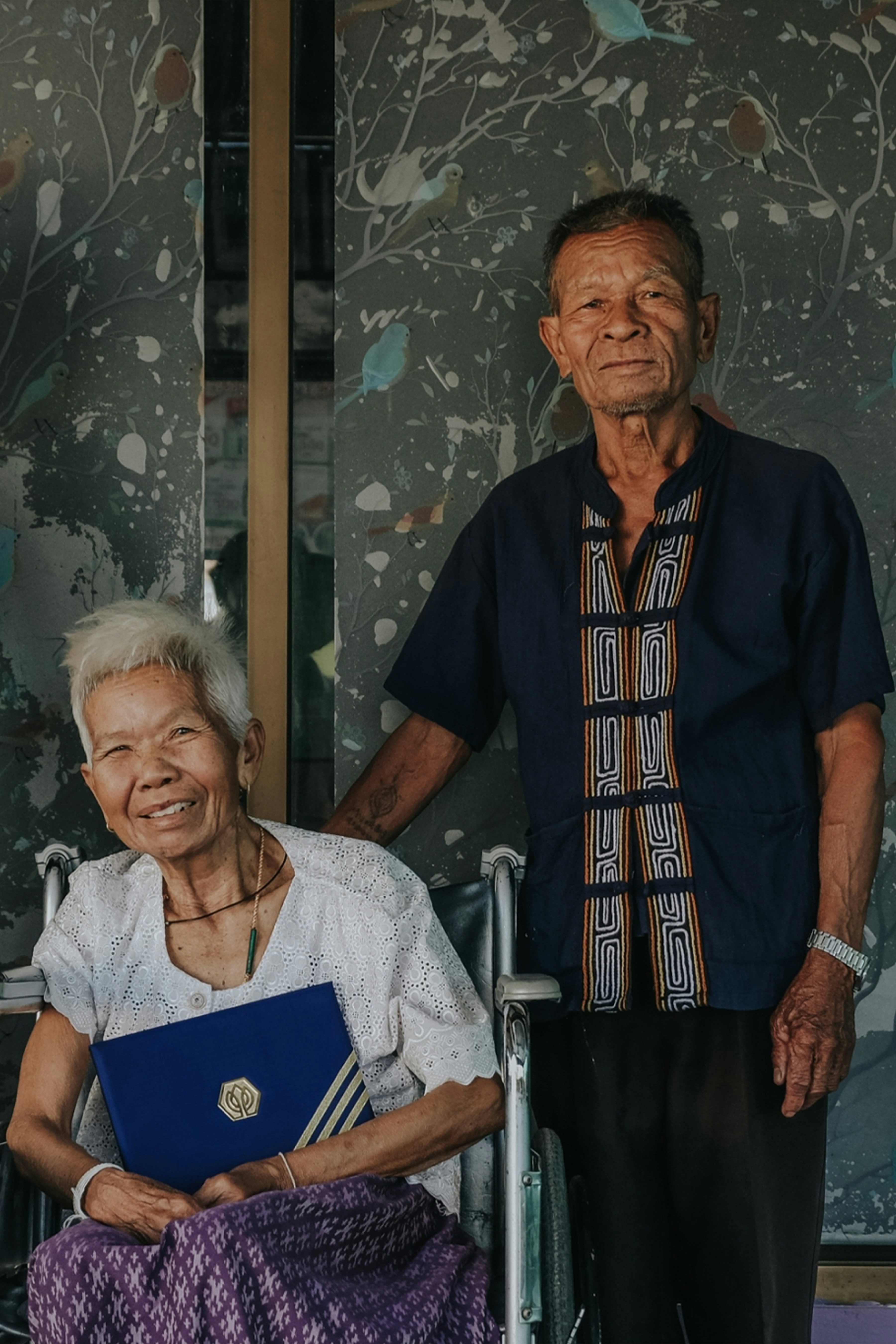 An elderly couple stands together, the woman seated in a wheelchair holding a blue folder with a gold logo. The man stands beside her, wearing a dark blue shirt with a patterned design. A decorative wall with a branching pattern and birds is in the background.
