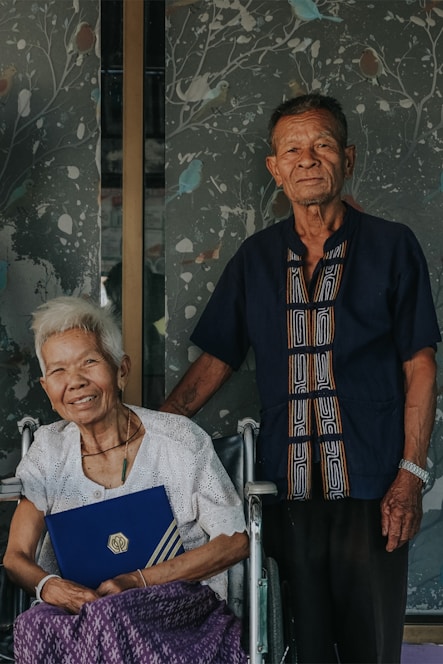 An elderly couple stands together, the woman seated in a wheelchair holding a blue folder with a gold logo. The man stands beside her, wearing a dark blue shirt with a patterned design. A decorative wall with a branching pattern and birds is in the background.