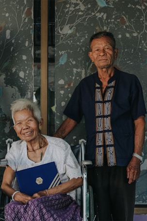 An elderly couple stands together, the woman seated in a wheelchair holding a blue folder with a gold logo. The man stands beside her, wearing a dark blue shirt with a patterned design. A decorative wall with a branching pattern and birds is in the background.