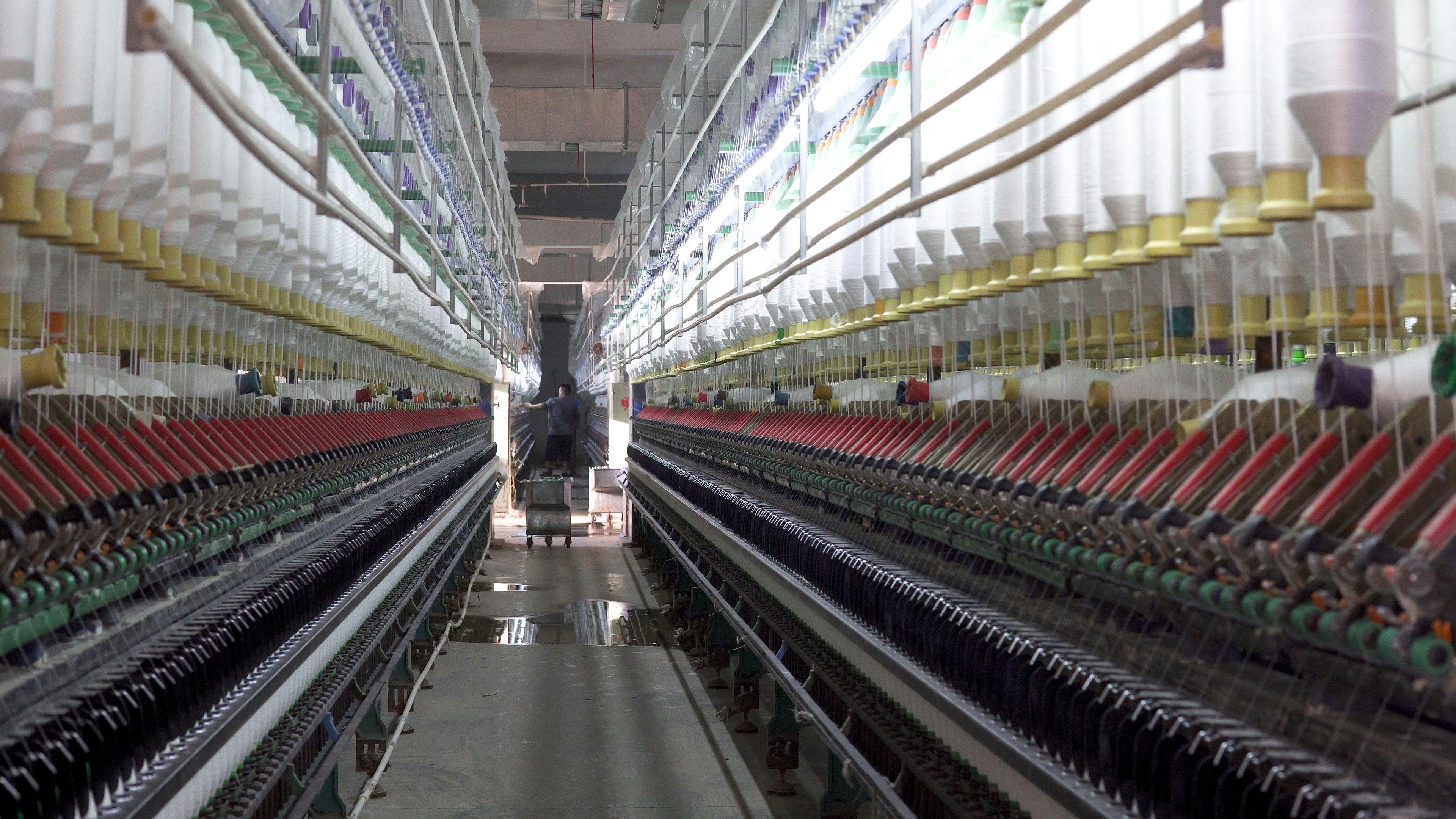 A symmetrical view down a corridor in a textile factory, flanked by rows of colorful spools of thread on both sides. The perspective draws the eye towards a worker and machinery at the far end, creating a sense of depth. The environment appears industrial and meticulously organized.