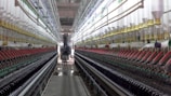 A symmetrical view down a corridor in a textile factory, flanked by rows of colorful spools of thread on both sides. The perspective draws the eye towards a worker and machinery at the far end, creating a sense of depth. The environment appears industrial and meticulously organized.