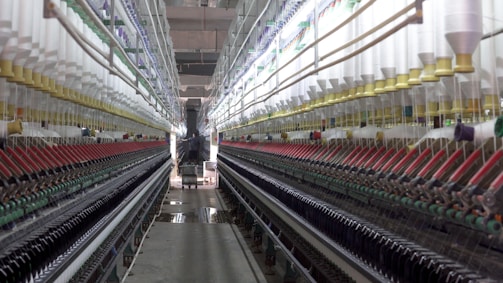 A symmetrical view down a corridor in a textile factory, flanked by rows of colorful spools of thread on both sides. The perspective draws the eye towards a worker and machinery at the far end, creating a sense of depth. The environment appears industrial and meticulously organized.