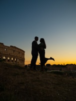 A silhouette of a couple embracing in front of a historic Roman structure during sunset. The sky is painted with warm hues transitioning to darker shades as night approaches.