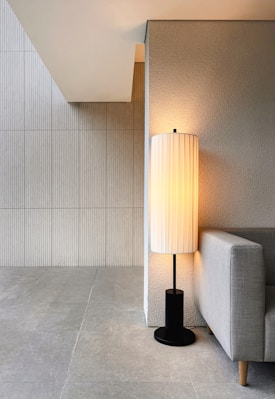 A modern, minimalist interior corner featuring a textured wall and tiled floor. A tall, cylindrical lamp with a pleated shade emits a warm glow, positioned beside a sleek gray sofa with wooden legs.