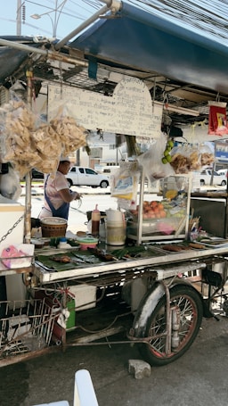 A traditional street food stall on a motorcycle cart, displaying snacks and fresh produce, covered by a blue tarpaulin. Various items are placed on banana leaves, and condiments are arranged on the counter. A vendor stands nearby, ready to serve customers. There are handwritten signs in a local language, and a bustling street with parked cars can be seen in the background.
