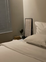 A neatly made bed with white sheets and pillows is placed in a corner room. To the side is a nightstand with a small potted plant, a wicker basket with a lid, and a framed text-based artwork leaning against the wall. The room has a simple, minimalist decor with closed blinds on the window next to the bed.