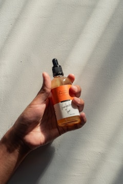 A hand holding a translucent bottle with an orange label and a dropper cap. The label has text, and the background is a textured, light-colored surface with soft shadows.