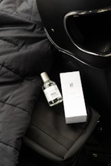 A black motorcycle helmet is situated on a textured black surface, next to a small bottle of perfume labeled 'HUE' and a corresponding white box labeled 'LAZ.' The items are placed on top of a black bag with a visible strap.