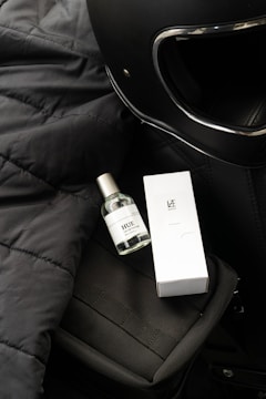 A black motorcycle helmet is situated on a textured black surface, next to a small bottle of perfume labeled 'HUE' and a corresponding white box labeled 'LAZ.' The items are placed on top of a black bag with a visible strap.