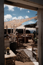An outdoor seating area with wooden furniture and potted plants is set beneath a woven canopy providing shade. The scene includes tables and chairs made of natural materials, a large potted plant with white branches, and a few people relaxing and enjoying the ambiance. The area is bathed in sunlight and is surrounded by a scenic view of distant hills and a bright blue sky peppered with fluffy clouds.