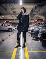 A person stands confidently in an underground parking garage, wearing a black outfit with a checkered scarf. The ceiling is lined with pipes and lights, and rows of parked cars are visible in the background.