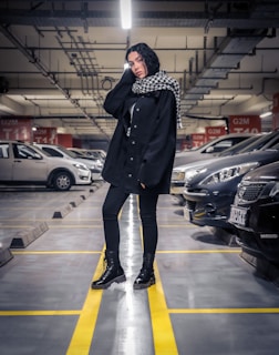 A person stands confidently in an underground parking garage, wearing a black outfit with a checkered scarf. The ceiling is lined with pipes and lights, and rows of parked cars are visible in the background.