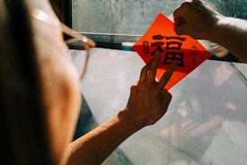 A person is delicately placing a red paper with Chinese characters on a window. The sunlight creates a warm reflection, and another hand visible in the image suggests an interaction or collaboration. The paper's vibrant color contrasts with the muted tones of the surrounding environment.