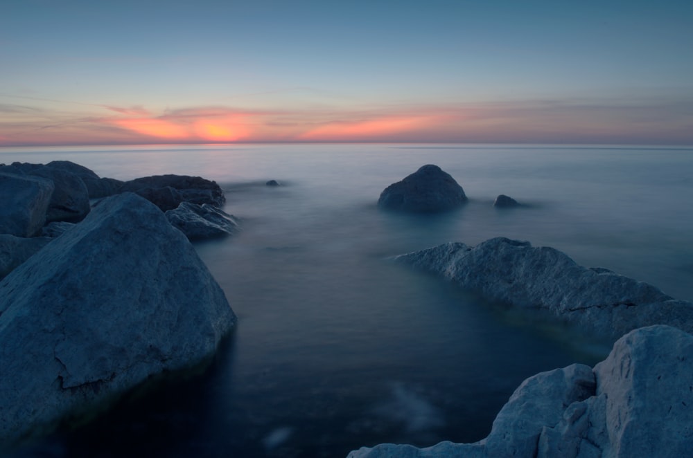 the sun is setting over the ocean with rocks in the foreground