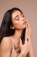 A woman with long, dark hair is shown with her eyes closed, gently touching her face with her hands. She wears a sleeveless, ribbed top in a soft brown shade. Her makeup is subtle and her nails are well-manicured.