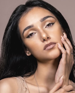 A close-up portrait of a woman with long dark hair and expressive eyes. She gently touches her face with her hand, showcasing a subtle, natural makeup look. The lighting highlights her facial features and gives her skin a soft glow.