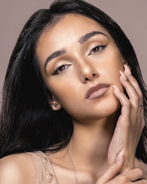 A close-up portrait of a woman with long dark hair and expressive eyes. She gently touches her face with her hand, showcasing a subtle, natural makeup look. The lighting highlights her facial features and gives her skin a soft glow.