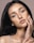 A close-up portrait of a woman with long dark hair and expressive eyes. She gently touches her face with her hand, showcasing a subtle, natural makeup look. The lighting highlights her facial features and gives her skin a soft glow.