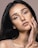 A close-up portrait of a woman with long dark hair and expressive eyes. She gently touches her face with her hand, showcasing a subtle, natural makeup look. The lighting highlights her facial features and gives her skin a soft glow.