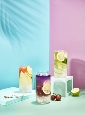 Three glasses of colorful beverages are artistically arranged, each filled with various fruits and ice cubes. The left glass contains a reddish liquid with orange and lemon slices. The middle glass displays a purple-hued drink with grapes and a lemon slice. The right glass holds a light green beverage with cucumber and lime slices. The backdrop features a pastel split between blue and pink tones, with a vibrant shadow of palm leaves cast on the left side. The setting includes pastel-colored geometric shapes and fruit pieces scattered around.