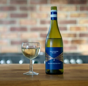 A bottle of Sauvignon Blanc labeled 'Brightwater Bay' stands on a wooden surface next to a partially filled glass of white wine. The background features a blurred brick wall, adding a rustic ambiance.