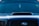 A close-up view of the front of a dark-colored car featuring a prominent vent on the hood. The windshield displays various decals or stickers, including a circular yellow one on the passenger side. Hanging from the rearview mirror is a small rectangular object.
