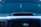 A close-up view of the front of a dark-colored car featuring a prominent vent on the hood. The windshield displays various decals or stickers, including a circular yellow one on the passenger side. Hanging from the rearview mirror is a small rectangular object.