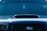 A close-up view of the front of a dark-colored car featuring a prominent vent on the hood. The windshield displays various decals or stickers, including a circular yellow one on the passenger side. Hanging from the rearview mirror is a small rectangular object.