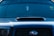 A close-up view of the front of a dark-colored car featuring a prominent vent on the hood. The windshield displays various decals or stickers, including a circular yellow one on the passenger side. Hanging from the rearview mirror is a small rectangular object.