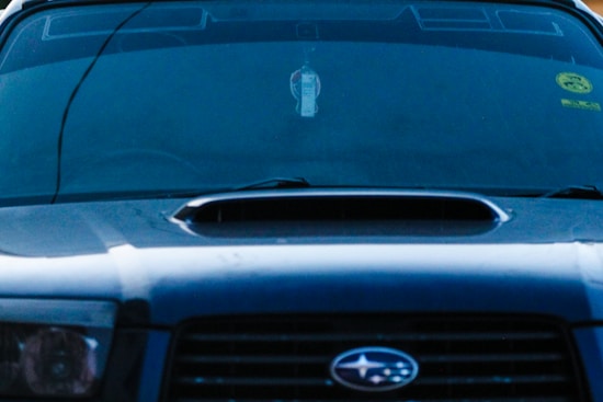 A close-up view of the front of a dark-colored car featuring a prominent vent on the hood. The windshield displays various decals or stickers, including a circular yellow one on the passenger side. Hanging from the rearview mirror is a small rectangular object.