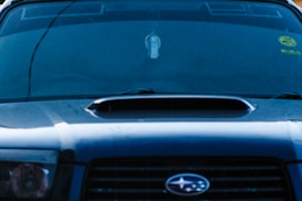 A close-up view of the front of a dark-colored car featuring a prominent vent on the hood. The windshield displays various decals or stickers, including a circular yellow one on the passenger side. Hanging from the rearview mirror is a small rectangular object.