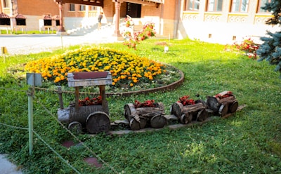 A miniature wooden train is creatively designed from logs, serving as a decorative garden feature. It sits on a grassy area, surrounded by vibrant yellow and red flowers within a circular flower bed. The background reveals part of a building with brick walls and decorative windows.