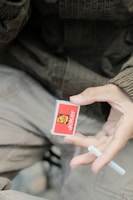 A hand holds a small box of Joker brand matches with a vibrant red design and a stylized joker face illustration. The person is also holding a single matchstick. The background features an out-of-focus, casual outfit with earthy tones.