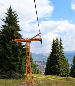 A ski lift with orange metal towers extends over a grassy hillside surrounded by tall evergreen trees. The lift cables stretch into the distance with chairlifts visible along the line. Puffy white clouds fill the blue sky, and rolling hills can be seen in the background.