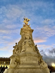 A grand monument featuring a statue with intricate sculptures, set against a vibrant sky displaying various shades of blue and orange. The structure is adorned with an ornate golden statue on top, and the base includes figures in classical poses.