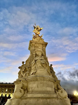 A grand monument featuring a statue with intricate sculptures, set against a vibrant sky displaying various shades of blue and orange. The structure is adorned with an ornate golden statue on top, and the base includes figures in classical poses.