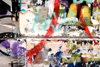 A colorful collage of peeling posters and graffiti on a wall, featuring layers of torn paper and various spray-painted shapes and colors. The artwork blends together, creating a chaotic yet visually engaging scene.